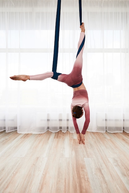 Mujer deportiva alegre vistiendo top y leggings realizando ejercicio de yoga antigravedad en el espacioso club de salud con ventanas panorámicas.