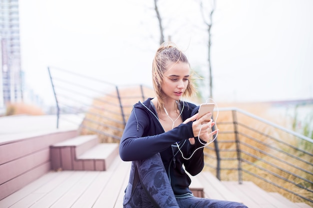 La mujer deportiva apta de los jóvenes que descansa y escucha música en el teléfono móvil después de entrenar