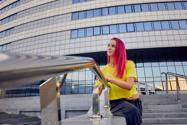 Mujer deportiva atlética con camiseta amarilla y cabello teñido de rosa está sentada junto al edificio del centro deportivo