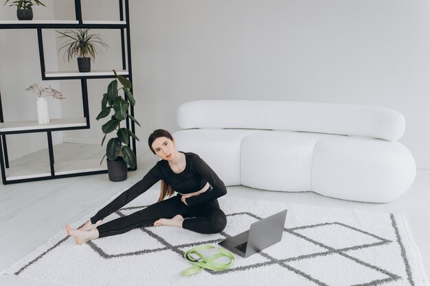 Mujer deportiva caucásica con ropa de fitness está en cuclillas usando una computadora portátil cerca de ella en la mañana