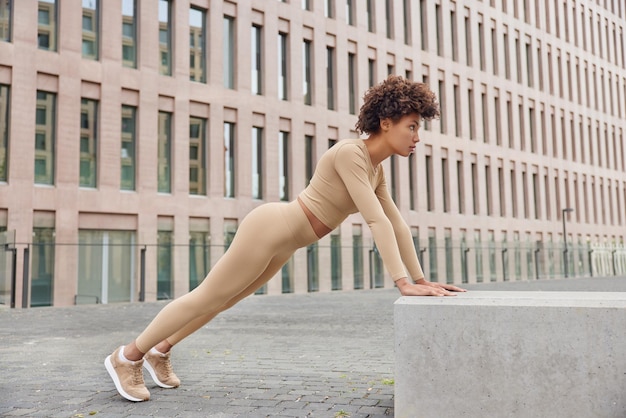 Mujer deportiva determinada hace ejercicios de flexiones al aire libre vestida con chándal y zapatillas de deporte posa al aire libre cerca de edificios urbanos tiene entrenamiento regular. Chica atlética muestra su fuerza entrena los músculos