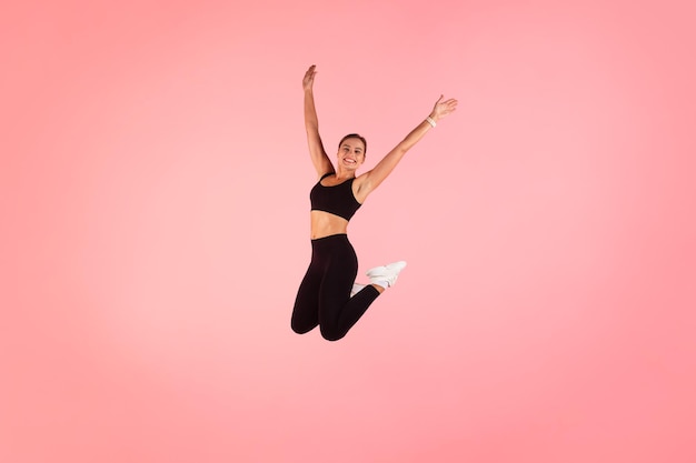 Mujer deportiva feliz en Fitwear saltando con las manos levantadas sobre fondo rosa