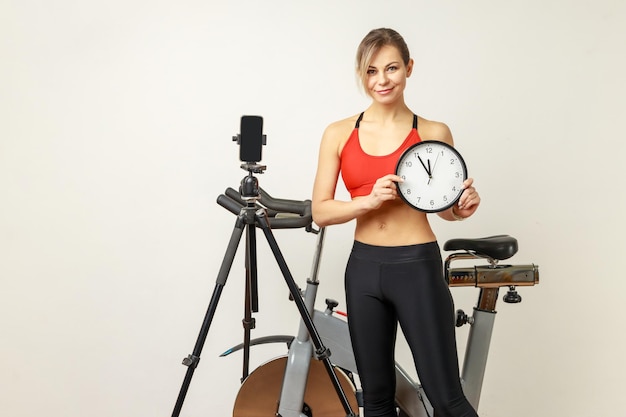 Mujer deportiva con forma perfecta de pie cerca del ejercicio y trípode con teléfono con reloj de pared