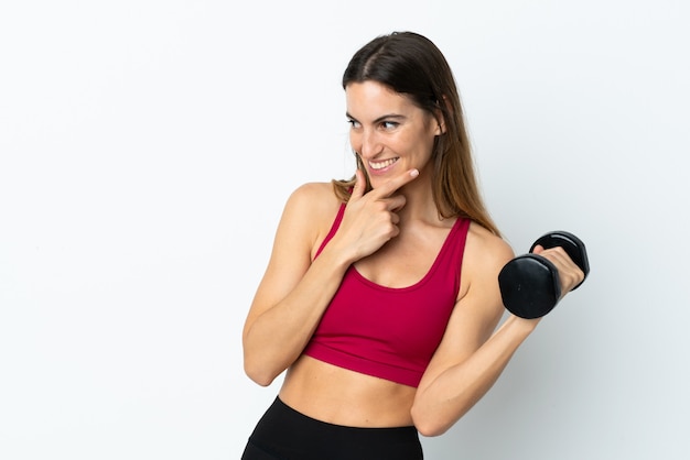 Mujer deportiva haciendo levantamiento de pesas en la pared blanca pensando una idea y mirando de lado