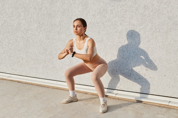 Mujer deportiva haciendo sentadillas de calentamiento, estirando cerca de una pared gris