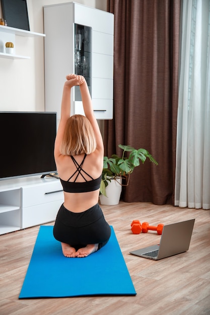 Mujer deportiva haciendo yoga fitness en casa a través de una computadora portátil por videollamada remota en línea. Mujer joven perdiendo peso por entrenamiento de gimnasio remoto en línea. Entrenamiento de la mujer irreconocible de la visión trasera.