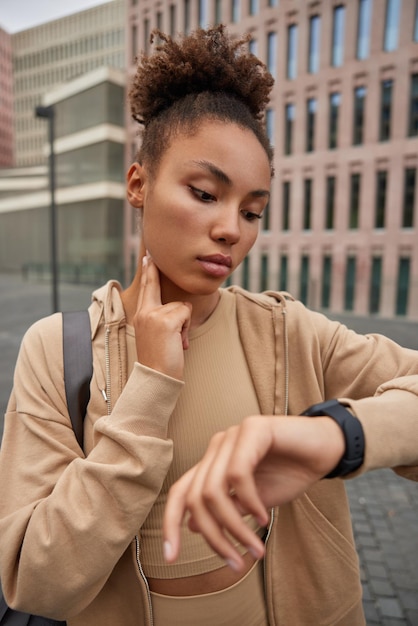 La mujer deportiva con el pelo rizado rastrea el pulso concentrado en el reloj inteligente mantiene la mano en el cuello monitorea el progreso vestida con poses de ropa deportiva contra la construcción. Estilo de vida fitness y concepto de salud