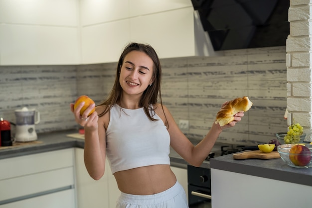 Mujer deportiva pensando qué comer en el desayuno entre frutas y bollo dulce en su cocina. Concepto de comida sana y no saludable