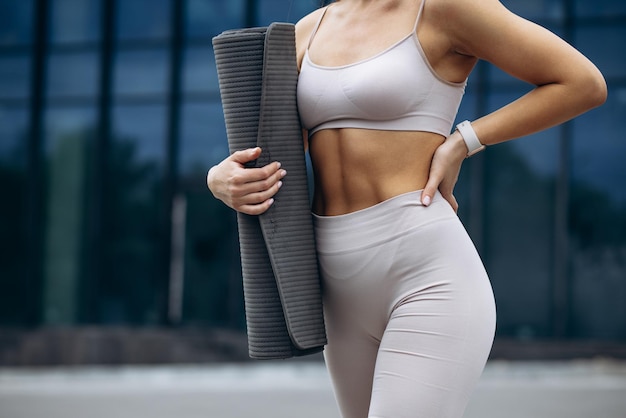 Mujer deportiva de pie con colchoneta de yoga junto al edificio