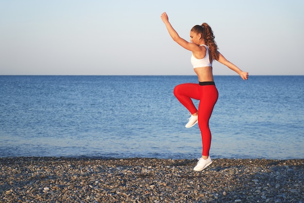 Mujer deportiva positiva en una mañana de verano entrenando en la playa en mallas rojas, entrenamiento en el fondo de la costa del mar, atleta saltando