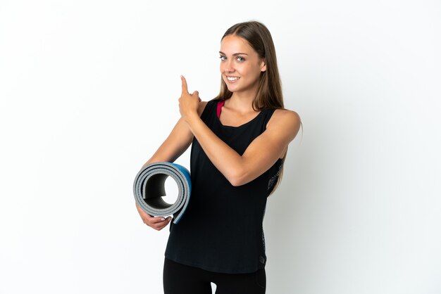 Mujer deportiva que va a clases de yoga mientras sostiene una estera sobre fondo blanco aislado apuntando hacia atrás