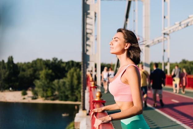 Mujer deportiva en ropa deportiva adecuada al atardecer en el puente moderno con vista al río mirar a su alrededor disfrutando del clima de verano