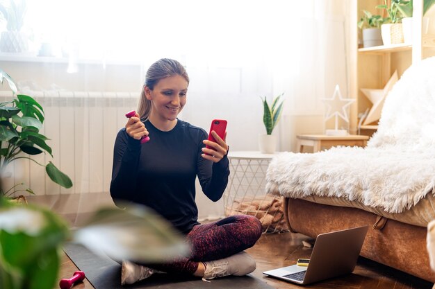 Una mujer deportiva en ropa deportiva y auriculares trabajando y usando la computadora portátil en casa en la sala de estar