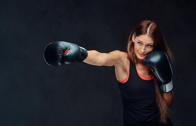 Mujer deportiva en ropa deportiva usando guantes de boxeo, entrenando en el gimnasio. Aislado en un fondo de textura oscura.