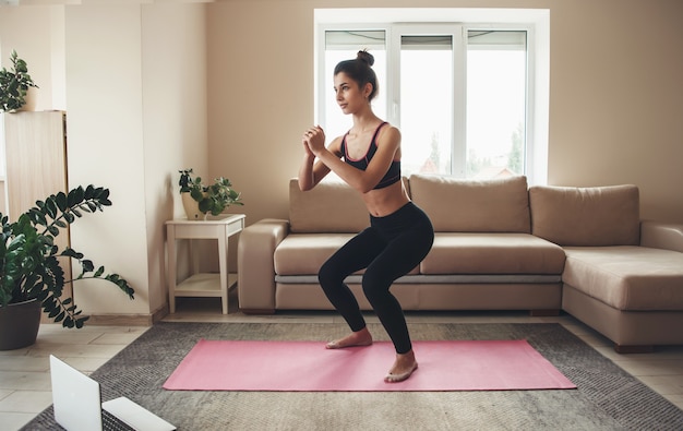 Mujer deportiva en ropa deportiva usando laptop y entrenando sus piernas en la alfombra de yoga en casa