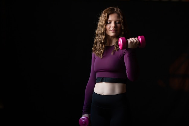 Mujer deportiva en ropa de entrenamiento con pequeñas pesas de color rosa en sus manos aisladas sobre fondo negro