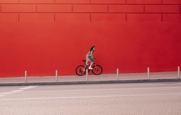 Mujer deportiva con ropa informal monta una bicicleta en una calle de la ciudad sobre el fondo de una pared roja