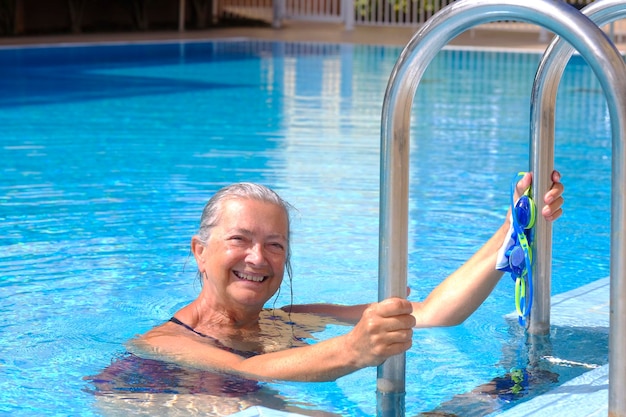 Mujer deportiva senior sonriendo fuera de la piscina con gafas de natación y gorra en la mano - jubilada activa disfrutando de nadar en un día soleado