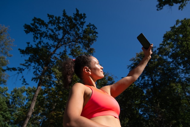 Foto mujer deportiva sosteniendo el teléfono móvil y haciendo selfie mientras se relaja después de hacer jogging al aire libre. disfrutando de la hermosa naturaleza