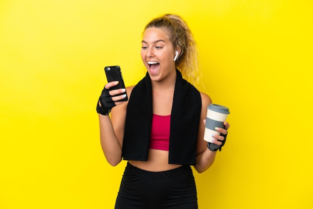 Mujer deportiva con toalla aislada de fondo amarillo sosteniendo café para llevar y un móvil