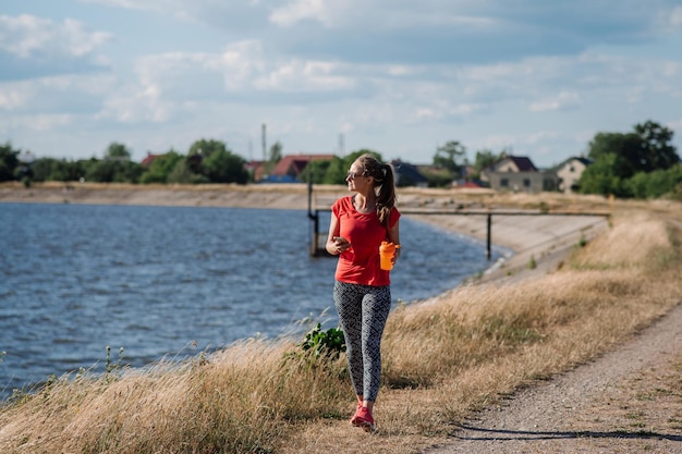 Mujer deportiva en traje deportivo caminando por el lago y usando un teléfono celular