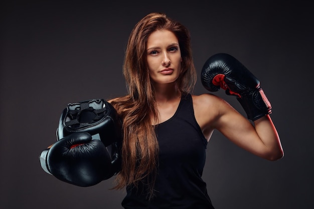 Mujer deportiva vestida con ropa deportiva con guantes de boxeo sostiene casco protector. Aislado en un fondo oscuro.