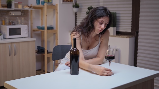 Mujer deprimida bebiendo una copa de vino sola en la cocina. Persona infeliz que sufre de migraña, depresión, enfermedad y ansiedad sintiéndose agotada con síntomas de mareos y problemas de alcoholismo.