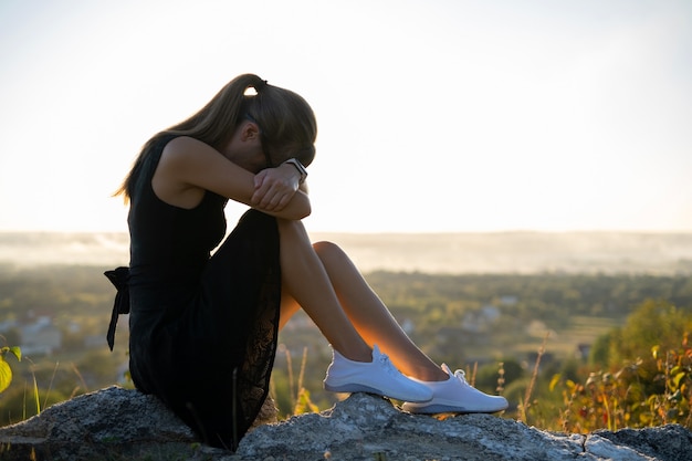 Mujer deprimida joven en vestido negro corto de verano sentado en una colina de la montaña.