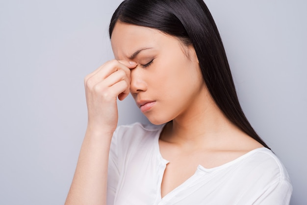 Mujer deprimida. Mujer asiática joven deprimida tocándose la cabeza con las manos y manteniendo los ojos cerrados mientras está de pie contra el fondo gris