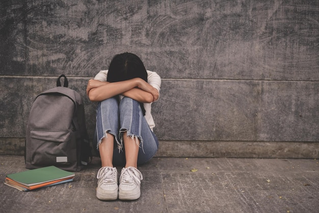 Mujer deprimida sentada con la mochila y los libros contra la pared en la acera