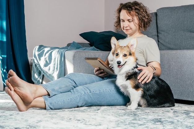 Mujer descalza alegre de pelo rizado con perro corgi sentado relajándose y leyendo un libro electrónico en la alfombra interior