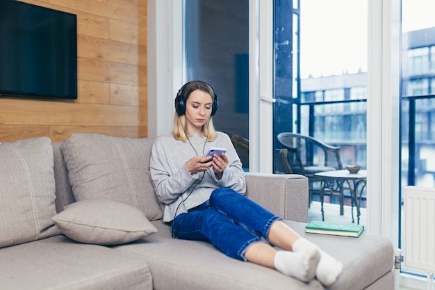 Mujer descansando en casa escuchando podcast de audio sentado en el sofá con auriculares