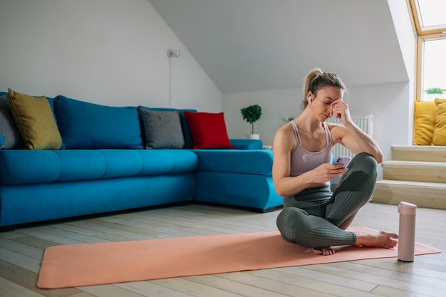 Mujer descansando después de hacer ejercicio de yoga en casa y usando un teléfono inteligente