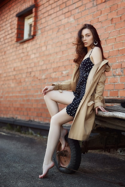 Mujer descansando en el pueblo cerca de los establos, retrato de una mujer en el sol, estilo rústico. Retrato de una morena sexy en un impermeable en la naturaleza. Maquillaje perfecto, cosmética natural.