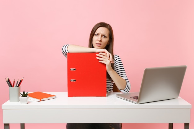 Mujer desconcertada apoyándose en la carpeta roja con documentos en papel y trabajando en un proyecto mientras está sentado en la oficina con un portátil