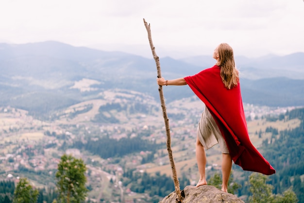 Mujer desconocida en vestido y capa roja de pie en piedra en la cima de la montaña. Mujer rubia descalza con gran palo de madera disfrutando del paisaje desde el punto de vista.