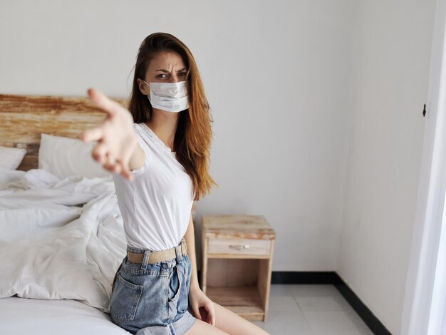 Foto mujer descontenta con mascarilla médica sentada sola en la cama en la sala de cuarentena coronavirus pandémico de vacaciones