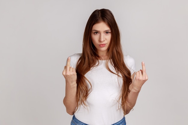 Mujer descortés y molesta que muestra el dedo medio a la cámara, expresa odio de protesta y falta de respeto, gesticulando con un símbolo grosero, usando una camiseta blanca. Disparo de estudio interior aislado sobre fondo gris.