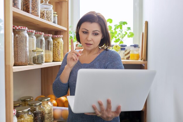 Mujer en la despensa de la cocina con productos almacenados con portátil