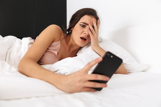 Mujer despierta mirando el teléfono móvil, mientras duerme en la cama sobre ropa blanca