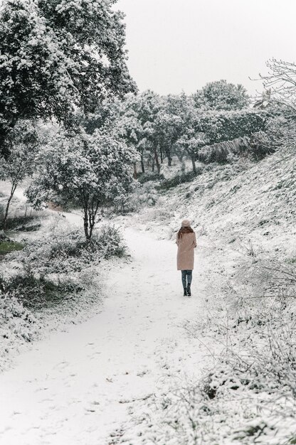 Mujer despreocupada en abrigo y sombrero caminando por el sendero en el bosque mientras disfruta de la naturaleza en invierno