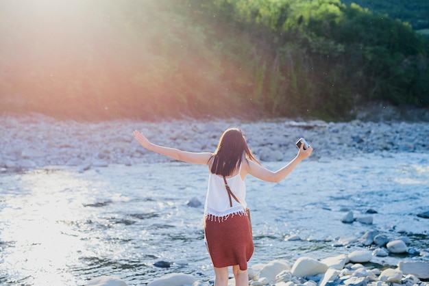 Mujer despreocupada caminando sobre el río para tomar fotos de la naturaleza con su cámara digital Mujer de vacaciones sola cruzando un río sobre rocas sosteniendo su cámara digital Mujer cruzando el río con una cámara digital