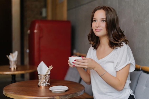 Mujer despreocupada con una camiseta blanca disfrutando de café aromático en un restaurante Expresión feliz mirada reflexiva en la distancia mientras pasa el tiempo libre en una mesa redonda de madera