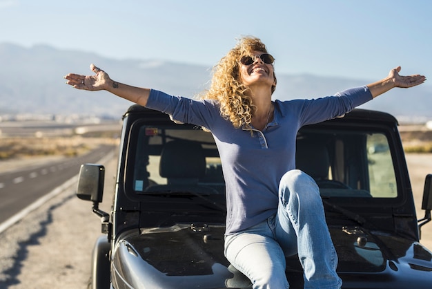 Mujer despreocupada sentada en el capó del coche aparcado en la carretera. Mujer joven alegre con los brazos extendidos disfrutando de sentarse en el capó del coche bajo el cielo