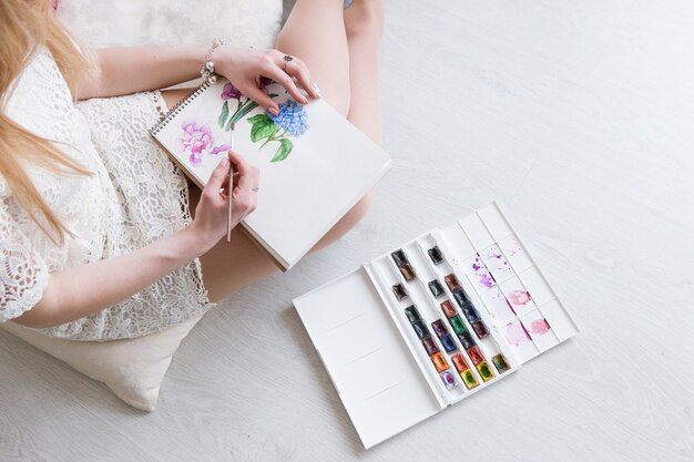 Mujer dibuja en acuarela vista superior Trabajo de pintor con boceto. Obras de arte coloridas de flores sobre papel blanco. Artista pinta con paleta y pincel.