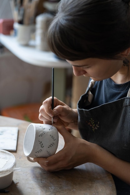 La mujer dibuja un patrón con pintura oscura en una taza de cerámica blanca usando un pincel en el fondo borroso