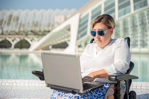 Mujer discapacitada concentrada sentada en silla de ruedas usa una computadora portátil al aire libre para trabajo remoto