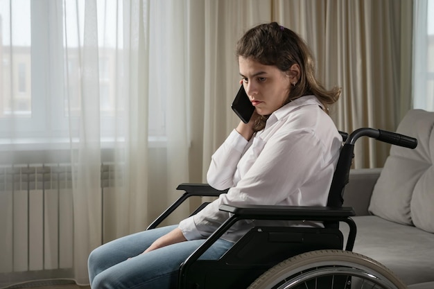 Mujer discapacitada con parálisis cerebral infantil habla sonriendo por teléfono móvil sentada en silla de ruedas cerca de la ventana en la sala de estar en la vista lateral de la casa