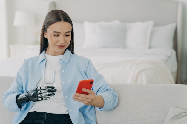 Mujer discapacitada con teléfono sosteniendo un vaso de agua con prótesis de brazo robótico en el dormitorio
