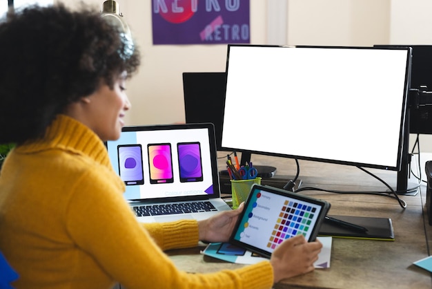 Mujer diseñadora afroamericana feliz usando tableta y computadora con espacio de copia en el escritorio en la oficina. Negocios creativos, diseño, tecnología y oficina casual.
